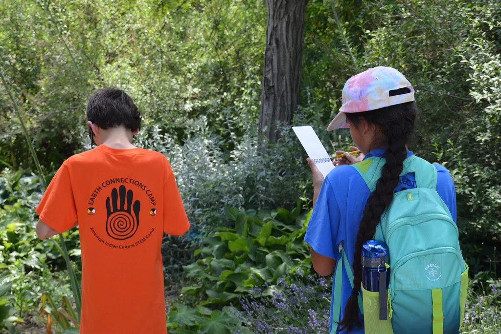 Two students seen from behind in a leafy garden, one in an orange camp shirt printed with a hand and spiral, the other with a long braid and a backpack writing on a notepad