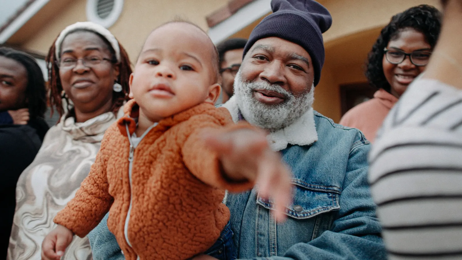 A smiling bearded man in a knit cap and denim jacket holds a baby in an orange fleece jacket who reaches toward the camera, with several women standing around them outside a house