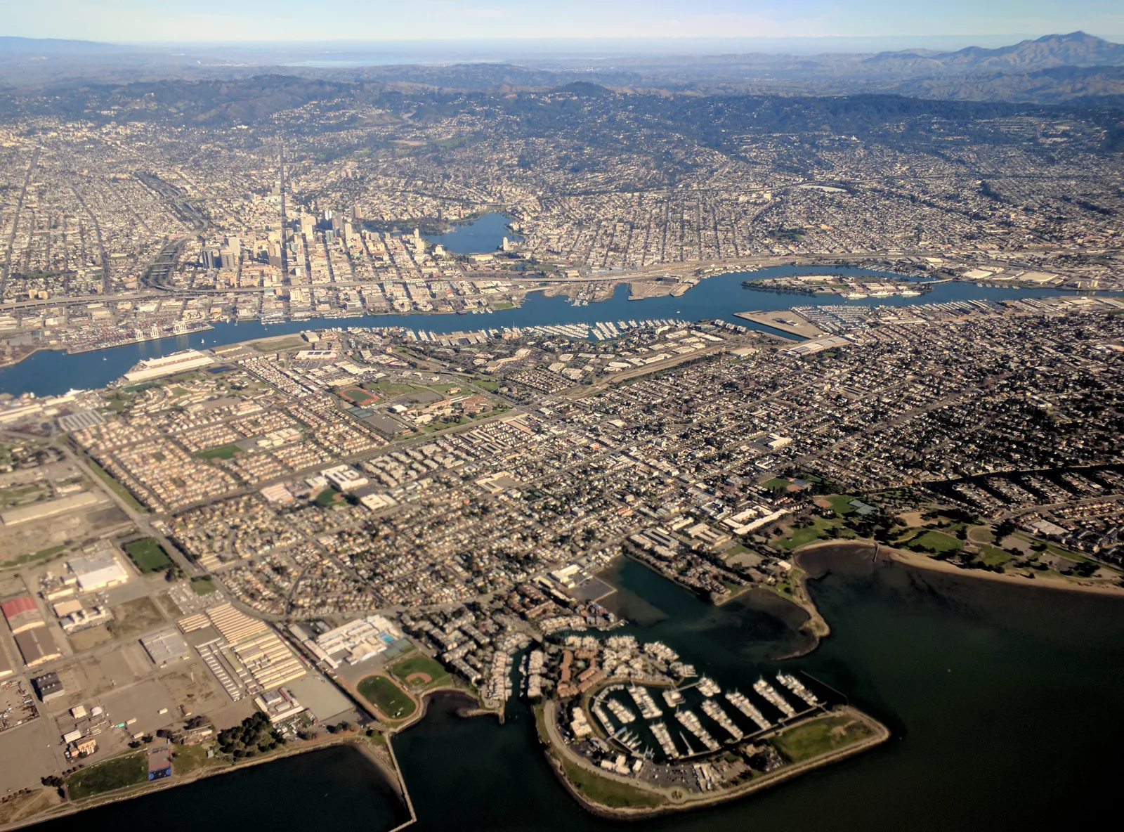 An aerial view over the city of Alameda, dense with houses and a marina in the foreground, across an estuary to the towers of downtown Oakland and Lake Merritt, with hills behind under a hazy sky