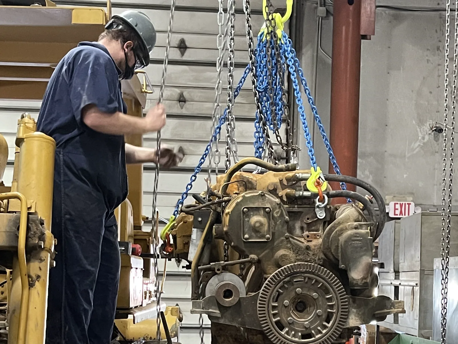 A worker in a hard hat and coveralls guides a diesel engine hanging from blue lifting chains in a heavy equipment repair shop