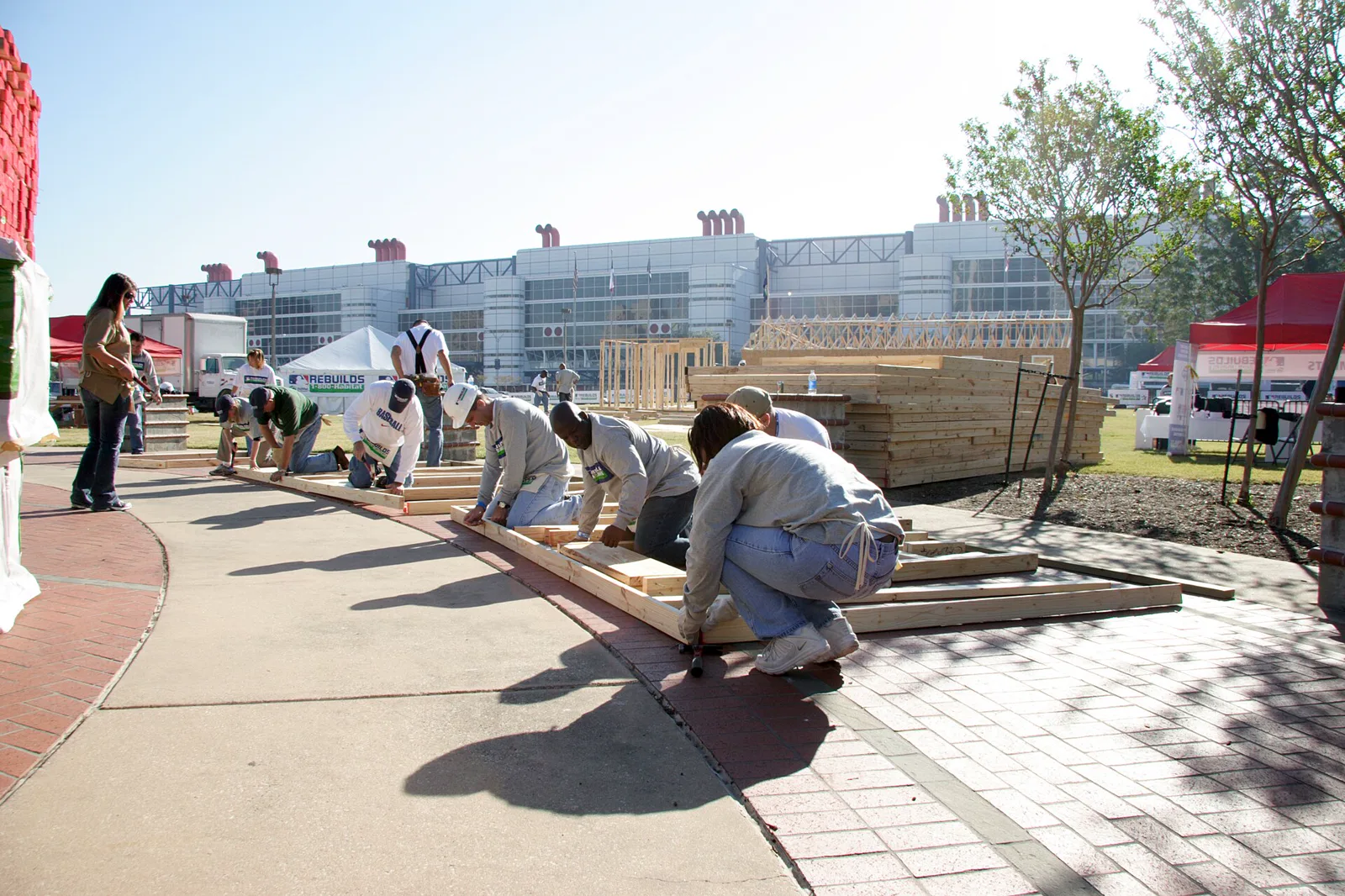 A row of volunteers in work clothes kneel on a paved walkway, bent over long wooden wall frames they are nailing together, with stacks of lumber, white tents and a large convention building behind them