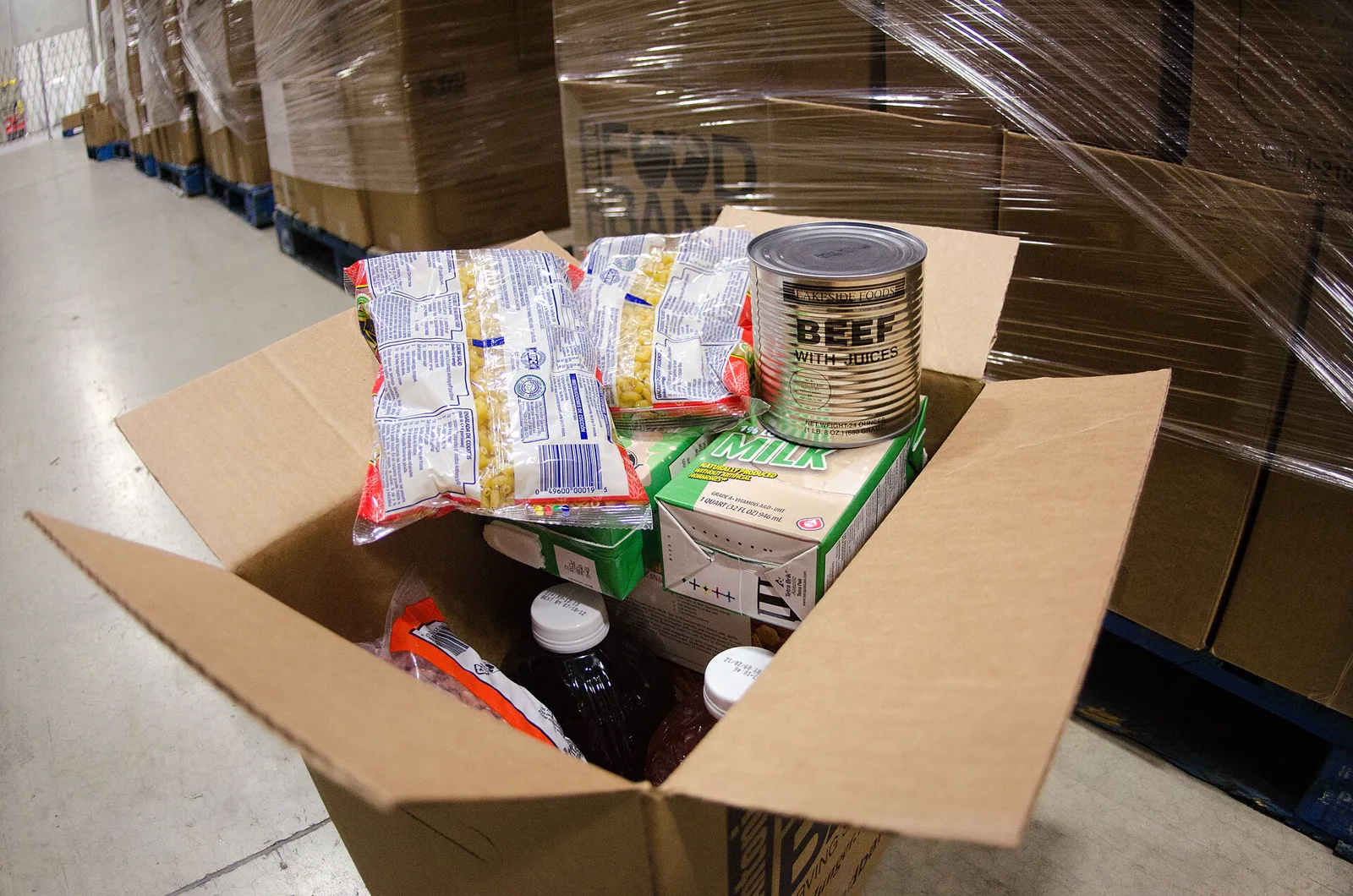 An open cardboard box of canned beef, pasta, boxed milk and juice on the floor of a food bank warehouse, beside pallets of boxes wrapped in plastic