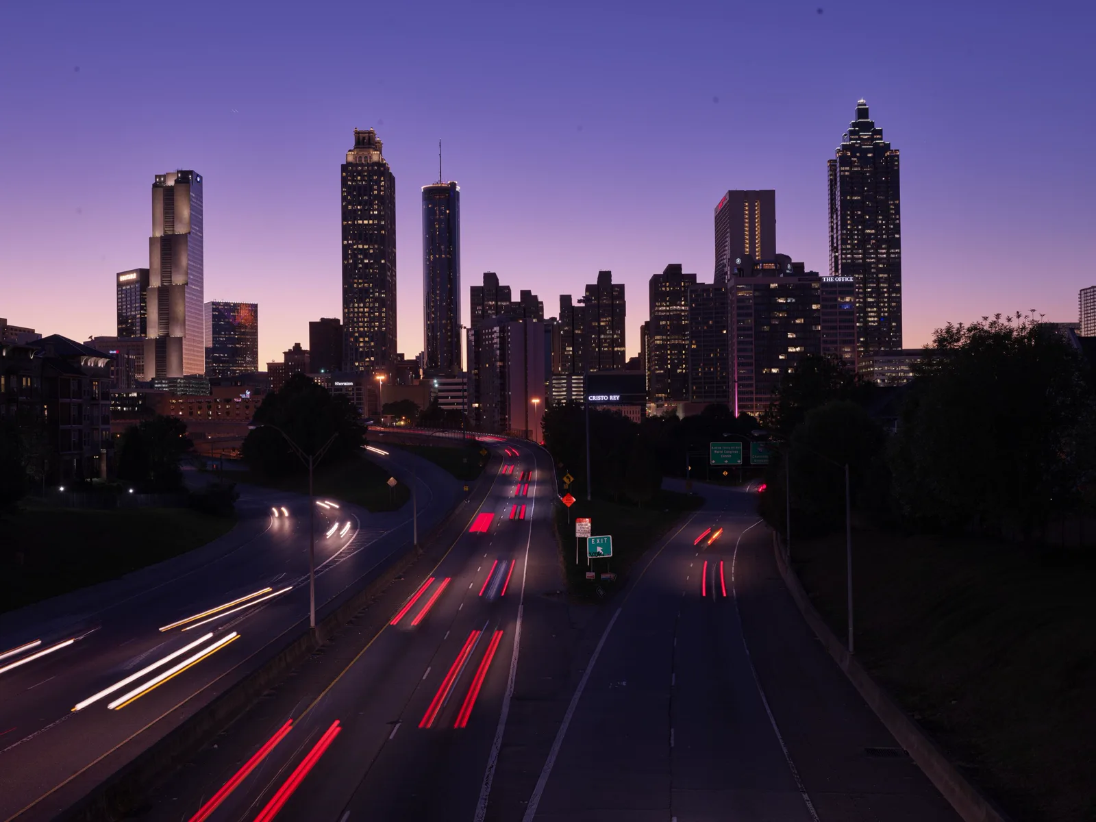 Lit office towers of downtown Atlanta against a purple dusk sky, above a curving highway streaked with the red and white light trails of passing cars