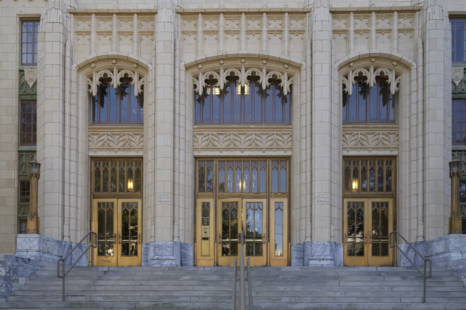 Three tall arched entrances with carved stone tracery above bronze and glass doors, the middle doors lettered City Hall, at the top of a wide flight of granite steps with metal handrails