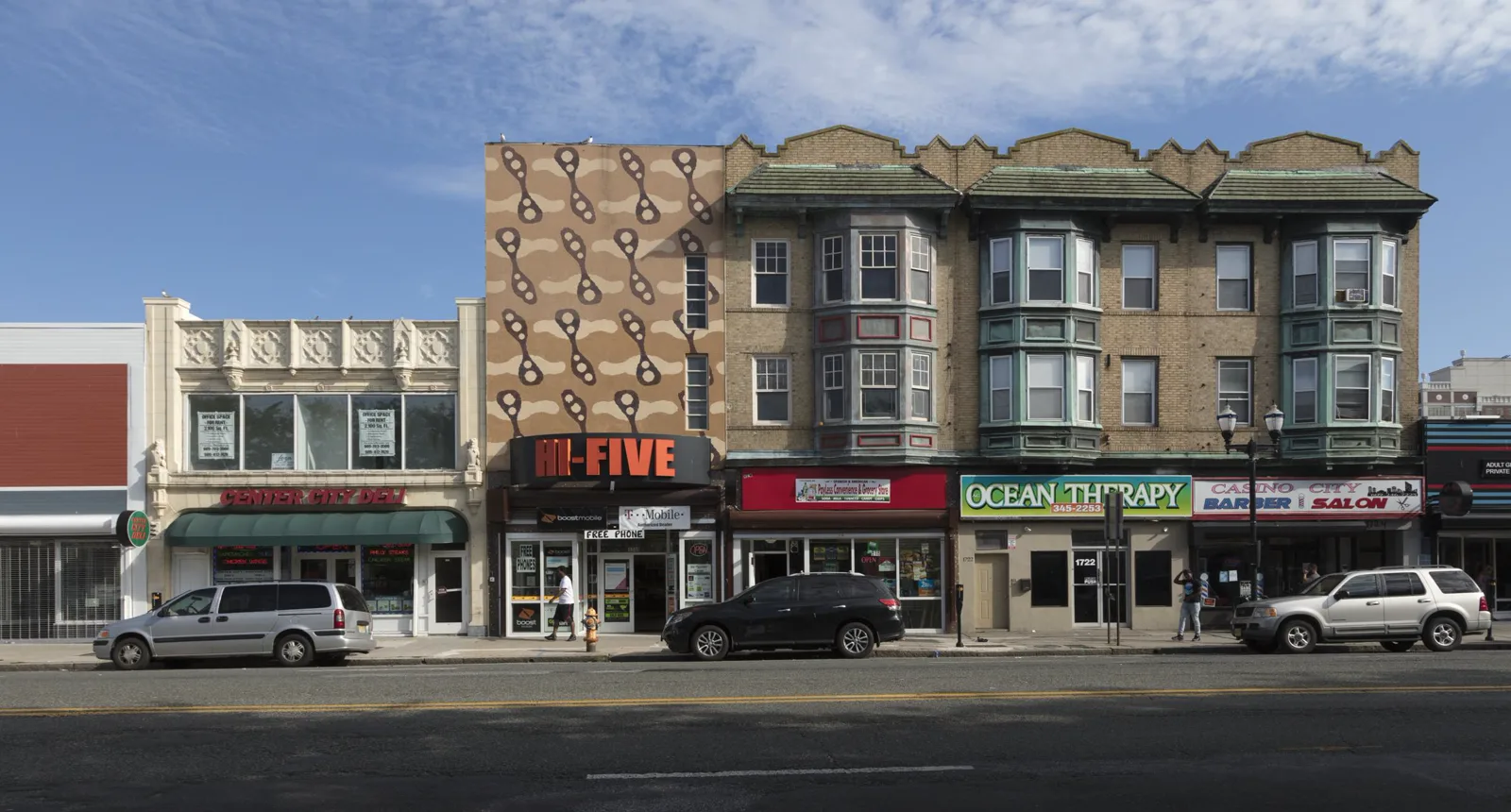 A row of three and four story brick buildings with shops on the ground floor, among them a barber and salon, along a wide street with parked cars under a blue sky