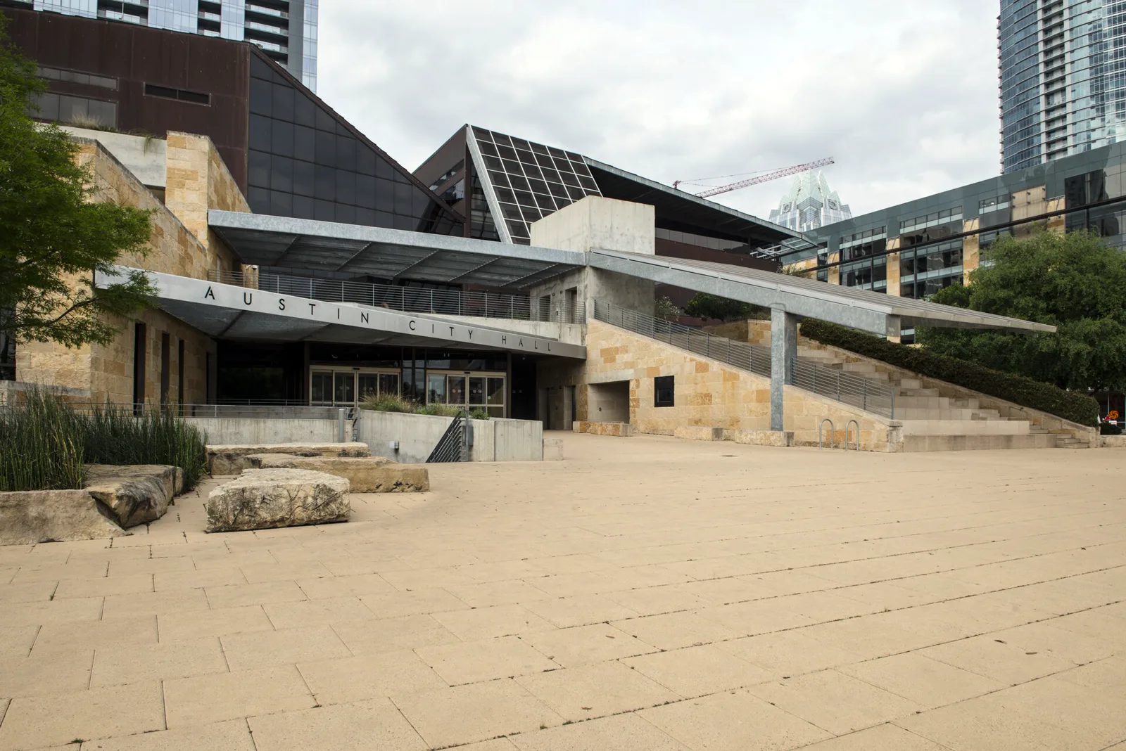 The entrance of a modern building of pale limestone, dark glass and steel, with the words Austin City Hall on a metal canopy, an empty paved plaza with limestone blocks in front and glass towers behind under a cloudy sky