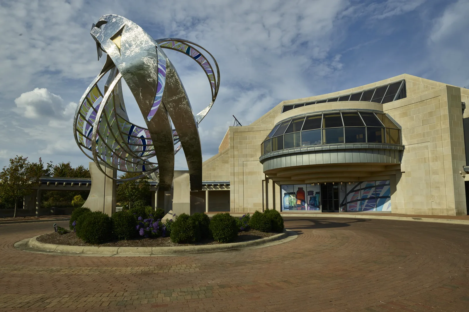 A tall stainless steel sculpture with colored glass panels stands on a planted traffic circle of red brick pavers in front of a limestone museum building with a curved glass front