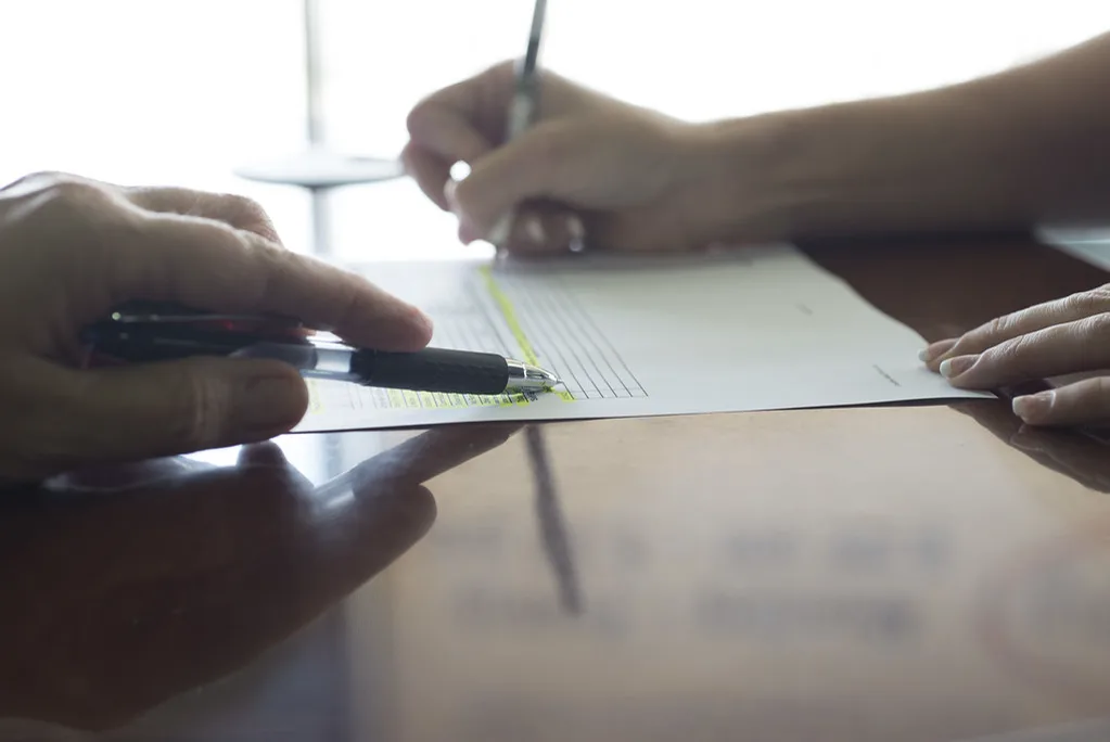 Close view of two people's hands over a form on a polished table, one holding a pen to the page while the other writes