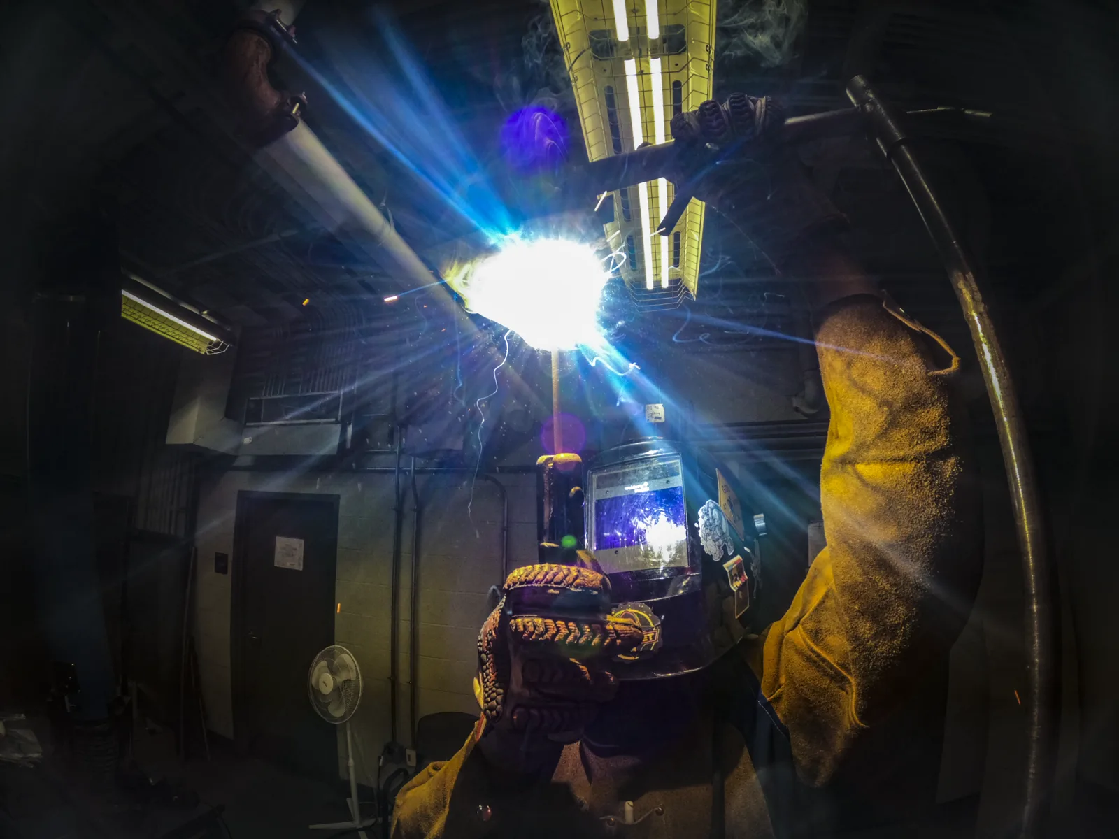 A welder in a helmet, leather jacket sleeve and heavy gloves holds a bright, sparking arc to a pipe overhead in a dim training shop