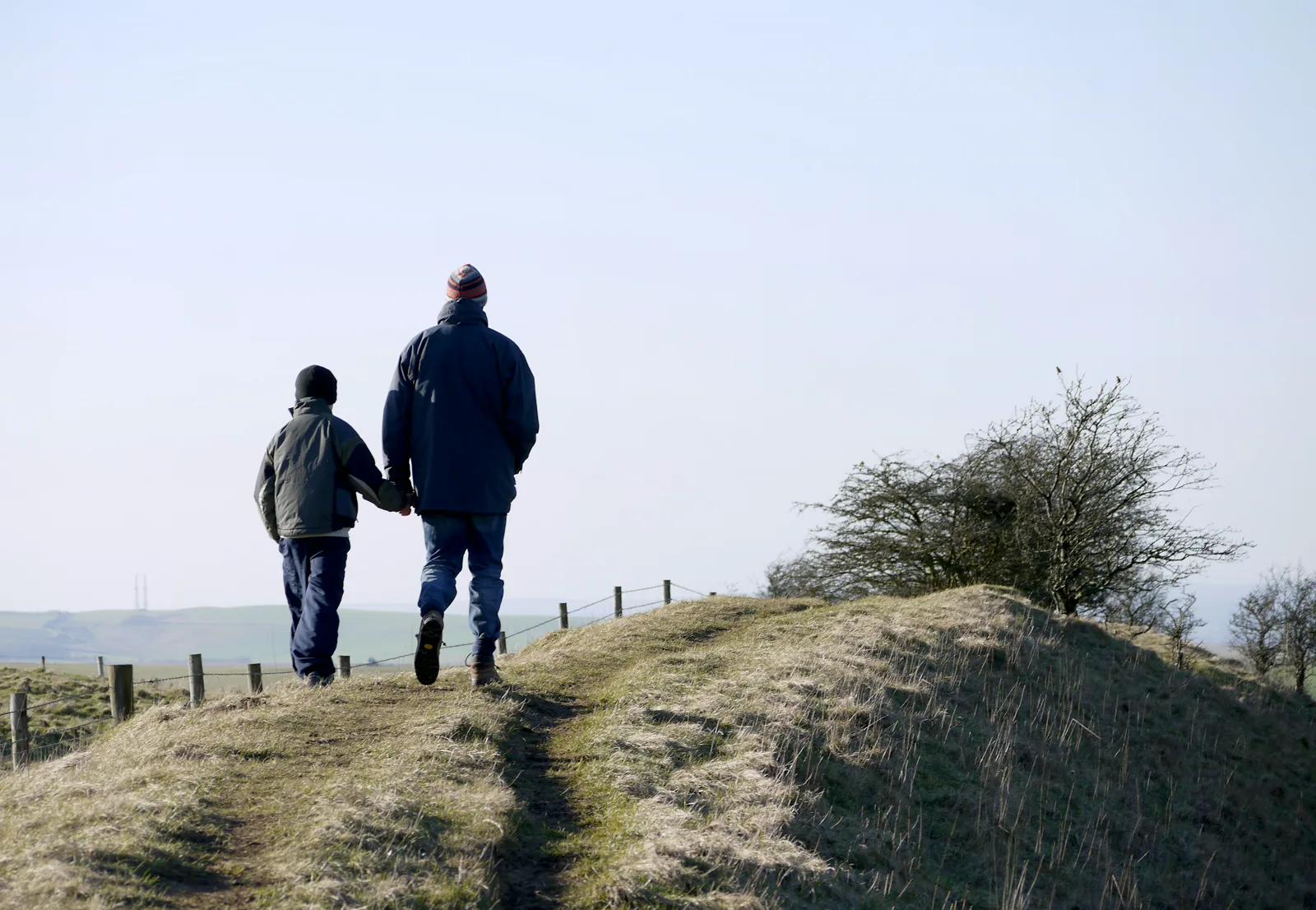 A man and a boy in winter coats and knit hats, seen from behind, walk hand in hand along a grassy ridge path beside a wire fence under a pale sky
