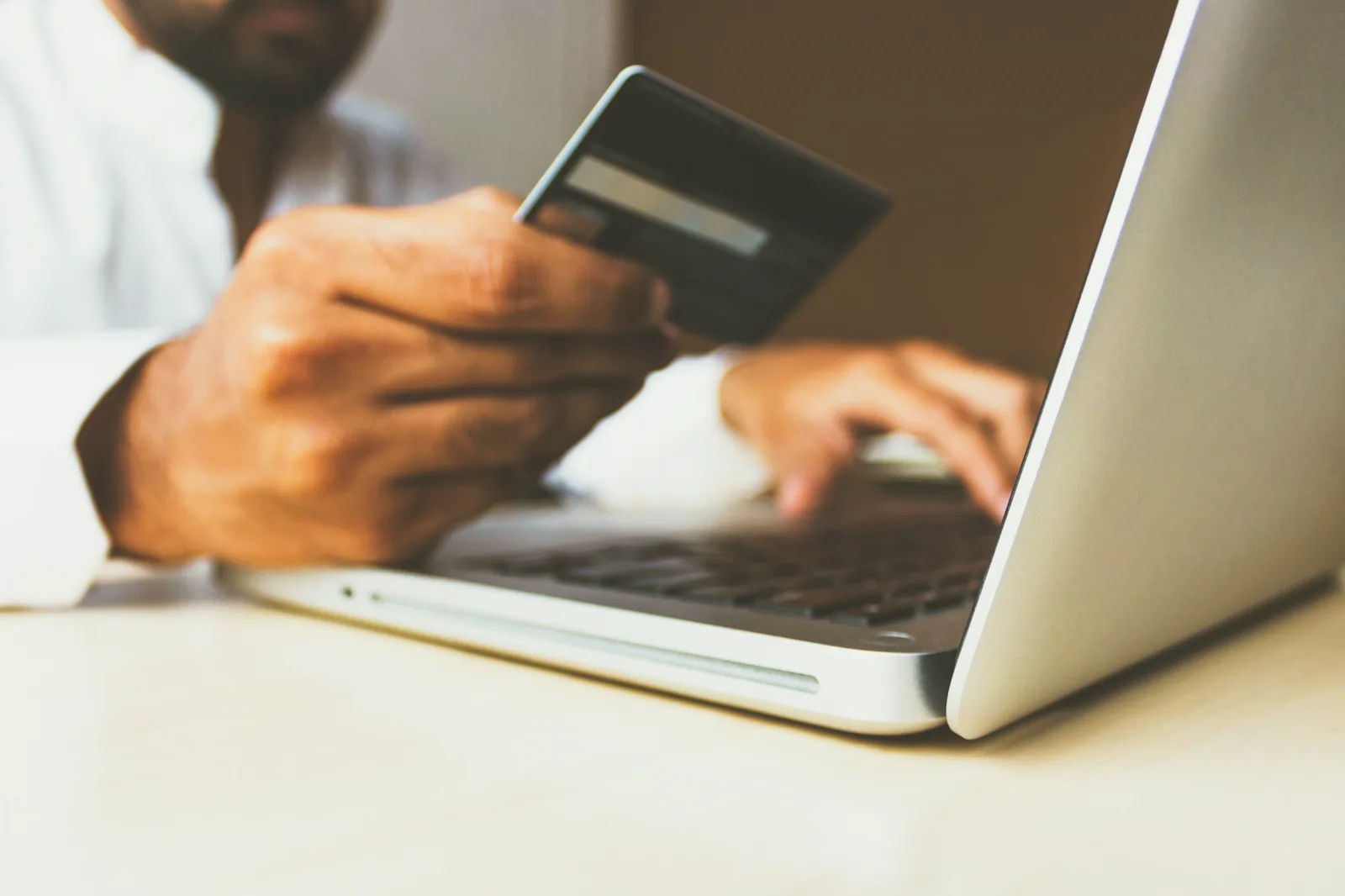 A hand holds a payment card beside an open silver laptop while the other hand types on the keyboard; the person's face is out of the frame