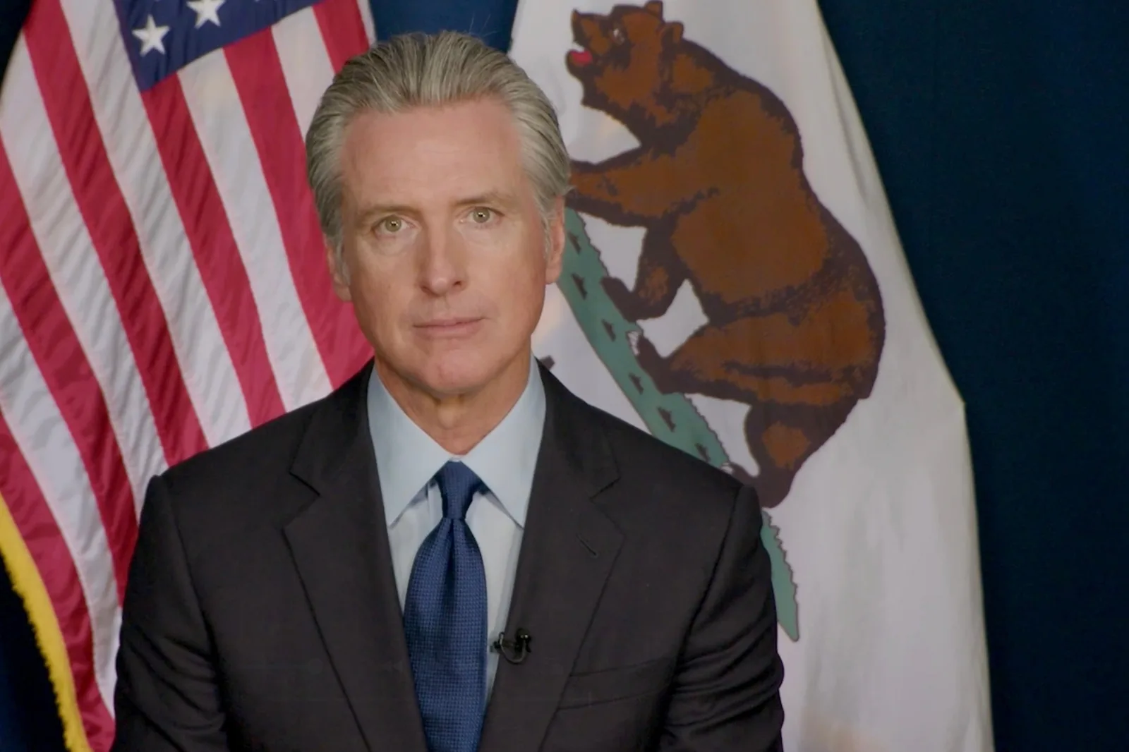 Gavin Newsom in a dark suit and blue tie stands in front of the United States flag and the California bear flag