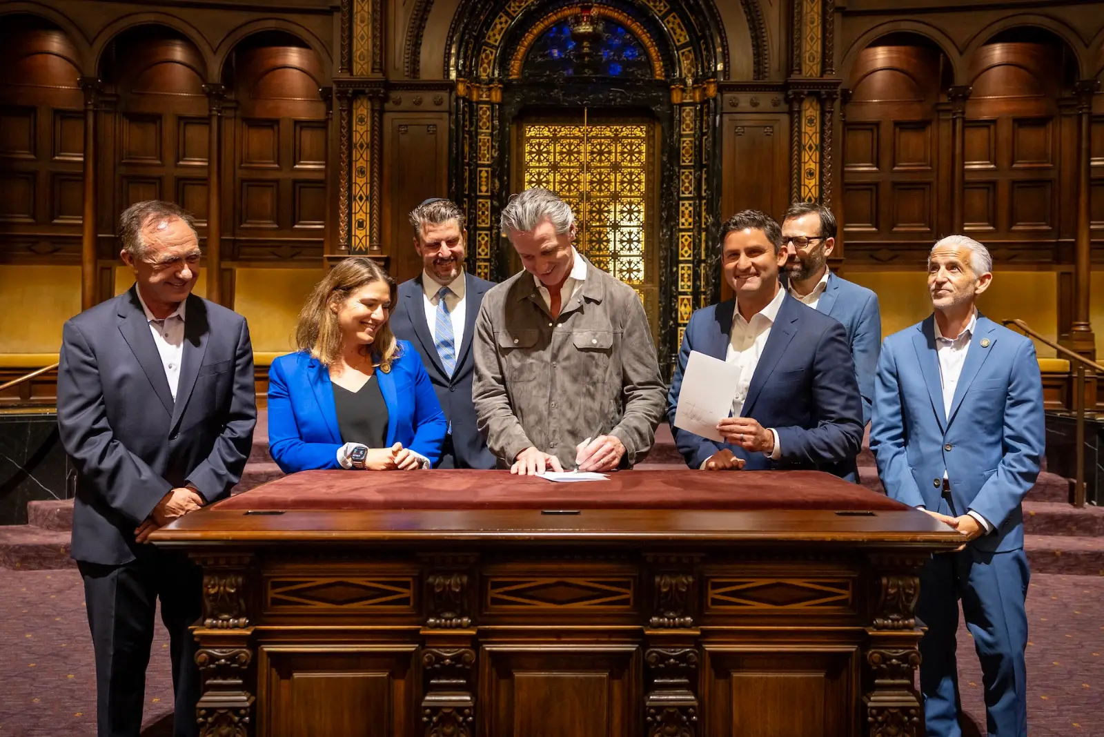 Gavin Newsom signs a document at a carved wooden desk in an ornate sanctuary while seven people stand around him