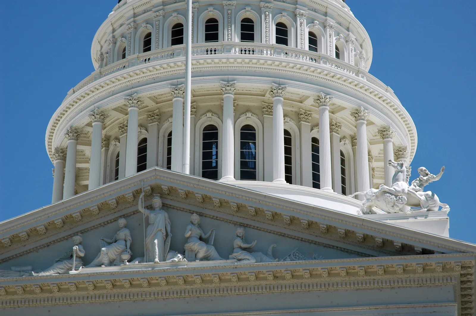 The white colonnaded dome of the California State Capitol rising behind a pediment filled with carved classical figures, against a deep blue sky