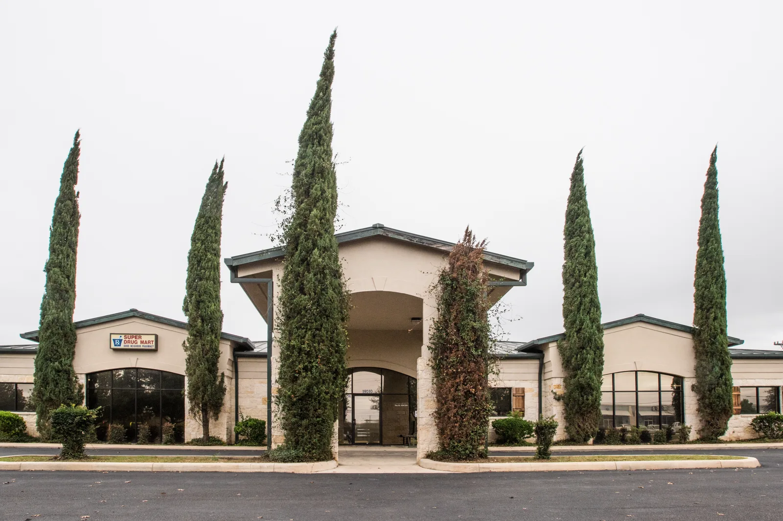 A single story beige stucco and stone clinic building with a peaked entrance canopy, framed by tall cypress trees, behind an empty parking lot under an overcast sky