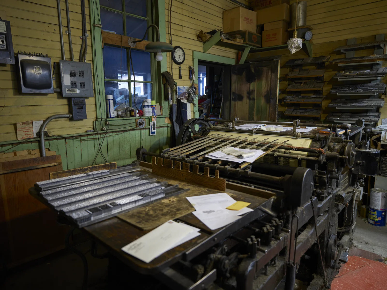 An old flatbed printing press in a cluttered wood-paneled shop, with rows of metal type locked in forms on a table in front, papers and envelopes on the press, and shelves of type cases along the back wall