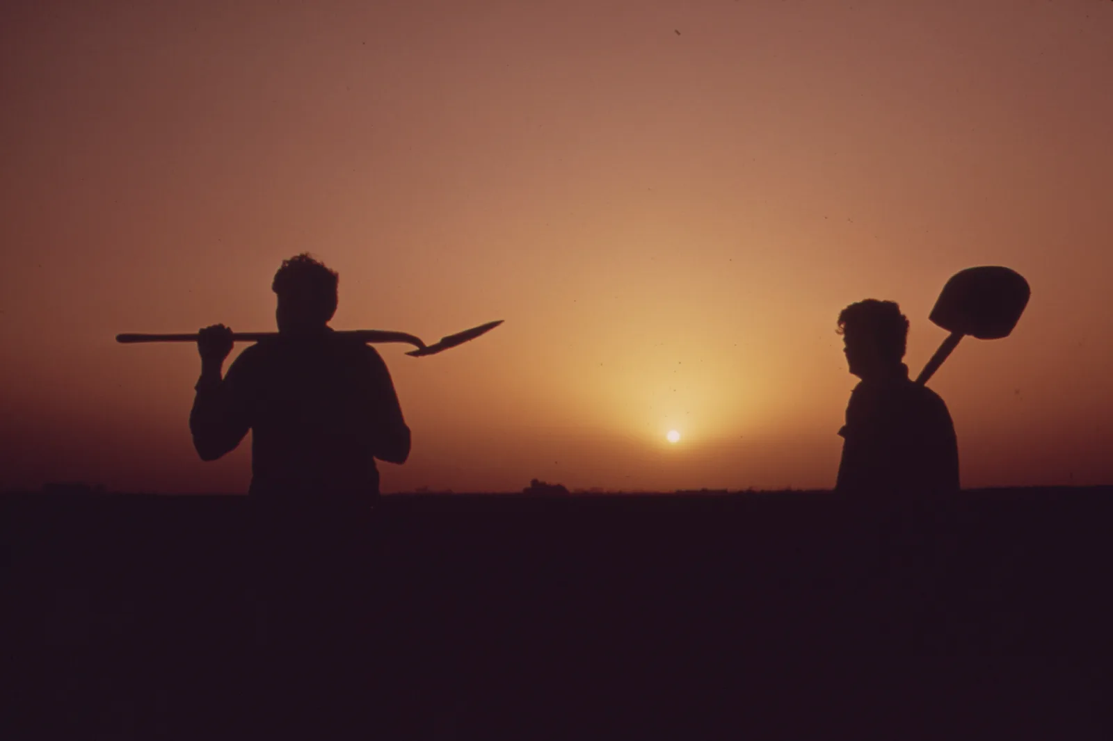 Two farm workers in silhouette against an orange sunset sky over flat fields, one carrying a shovel across his shoulders and the other a shovel over one shoulder