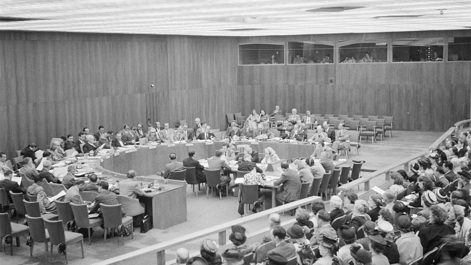 Black and white photo of delegates seated at a curved horseshoe table in a wood-paneled meeting room, with a public gallery of spectators in the foreground and interpreters' booths above
