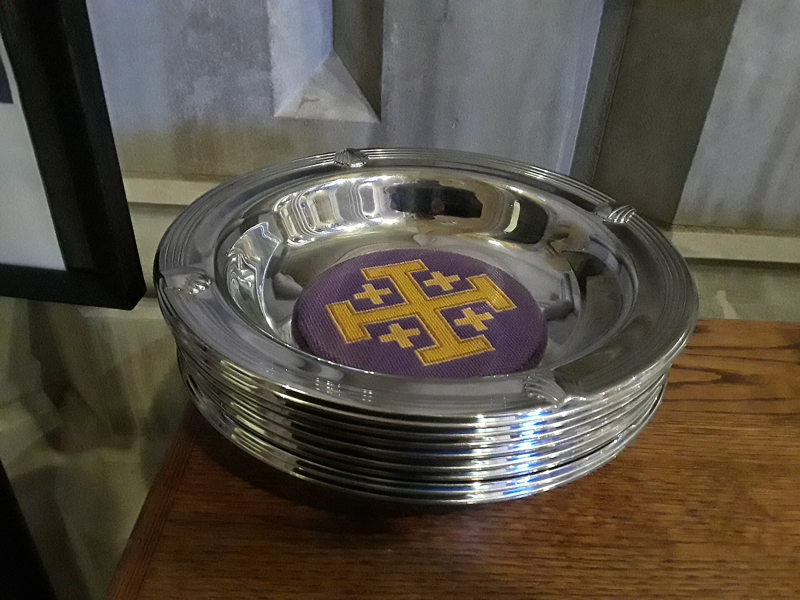 A stack of polished silver collection plates on a wooden table, the top one lined with a purple needlepoint cushion bearing a gold cross