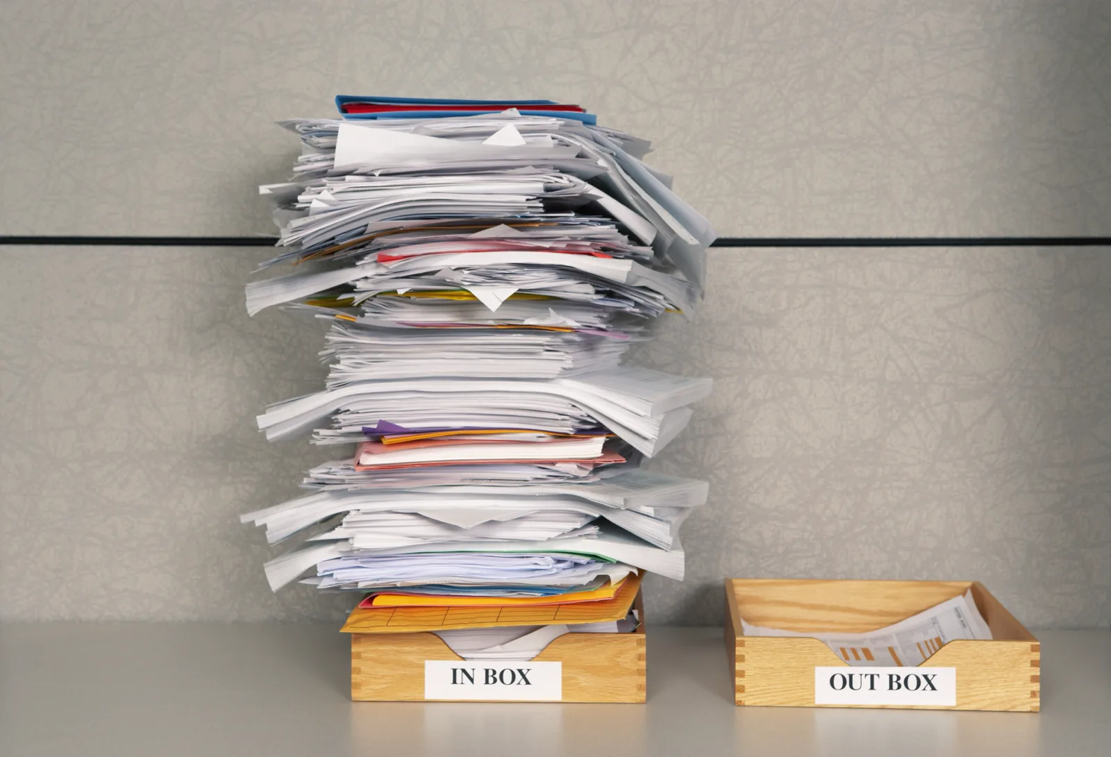 A tall, uneven stack of papers and folders in a wooden tray labeled In Box, beside a second tray labeled Out Box that holds only a few sheets, against a gray office partition