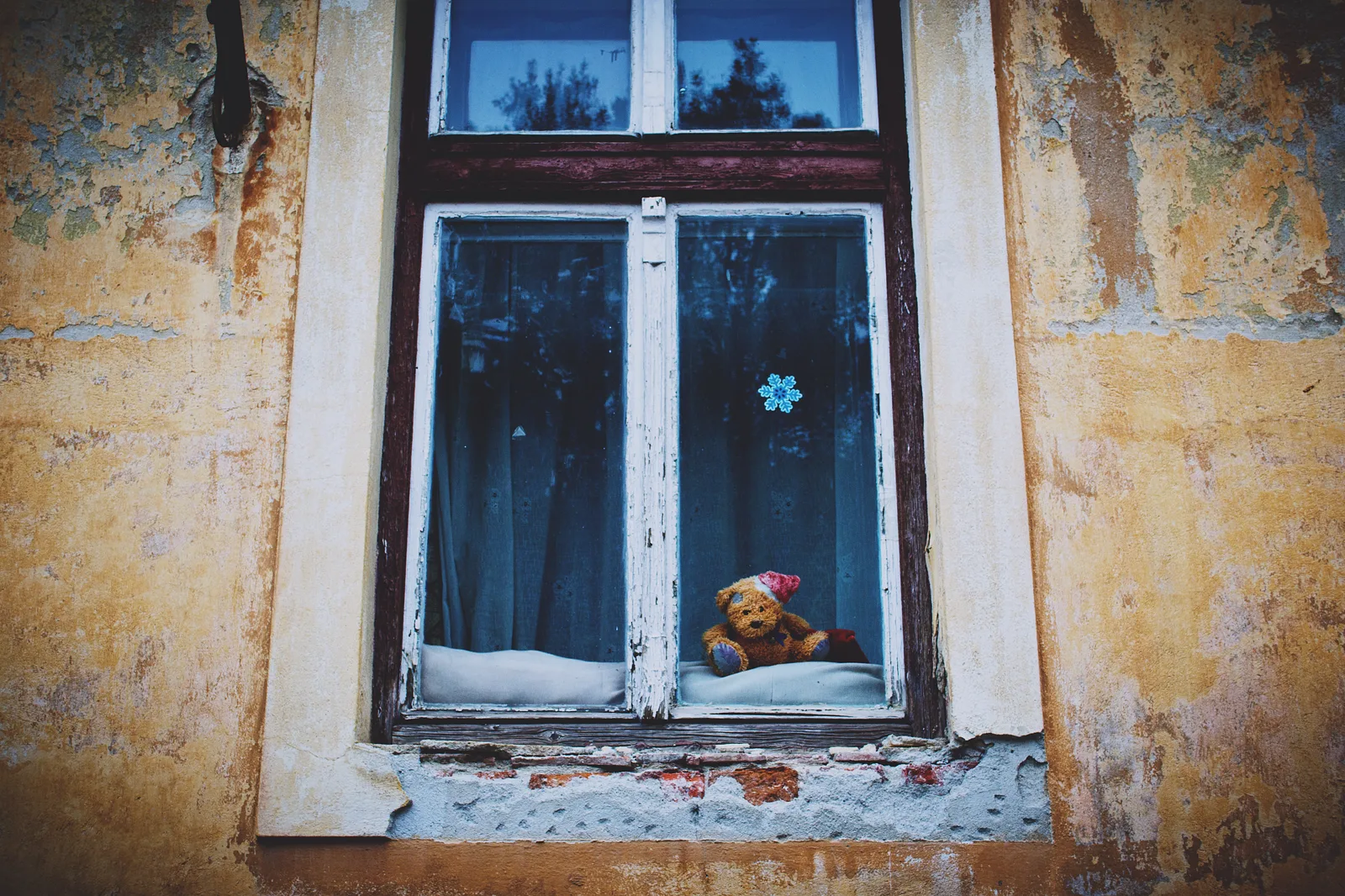 A teddy bear in a red knitted cap sits on the sill inside an old window with peeling paint, set in a weathered yellow wall