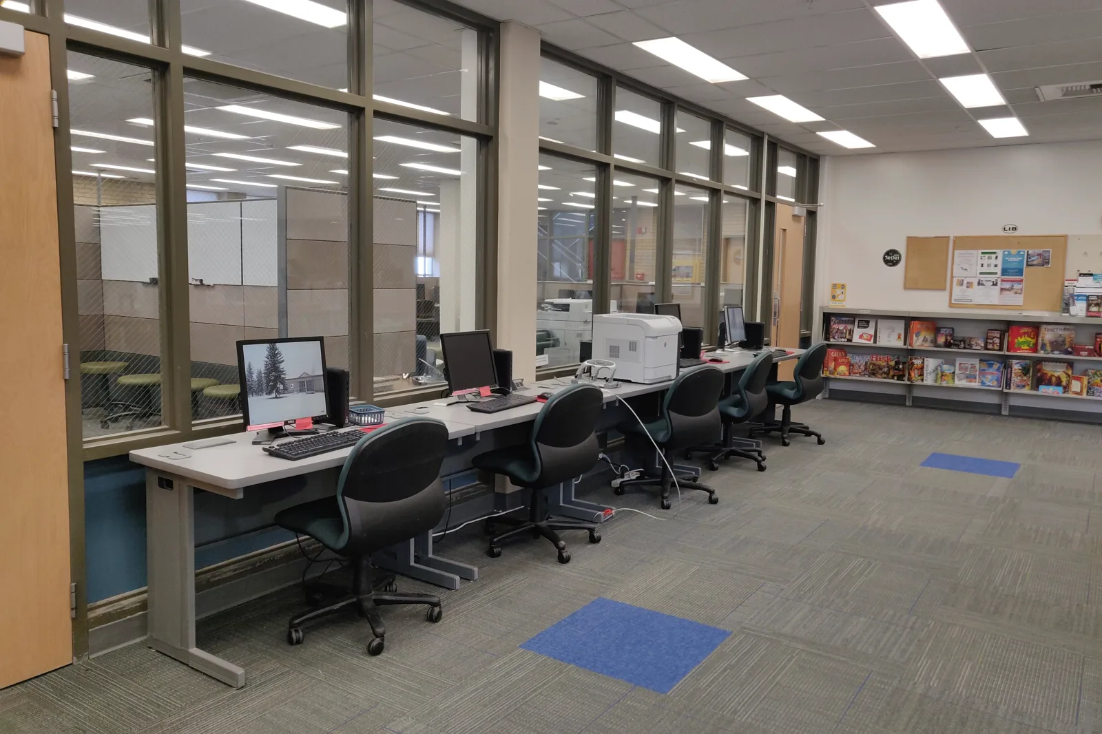 A row of desktop computers, a printer and empty office chairs along a window wall in a library, with board games on low shelves at the right