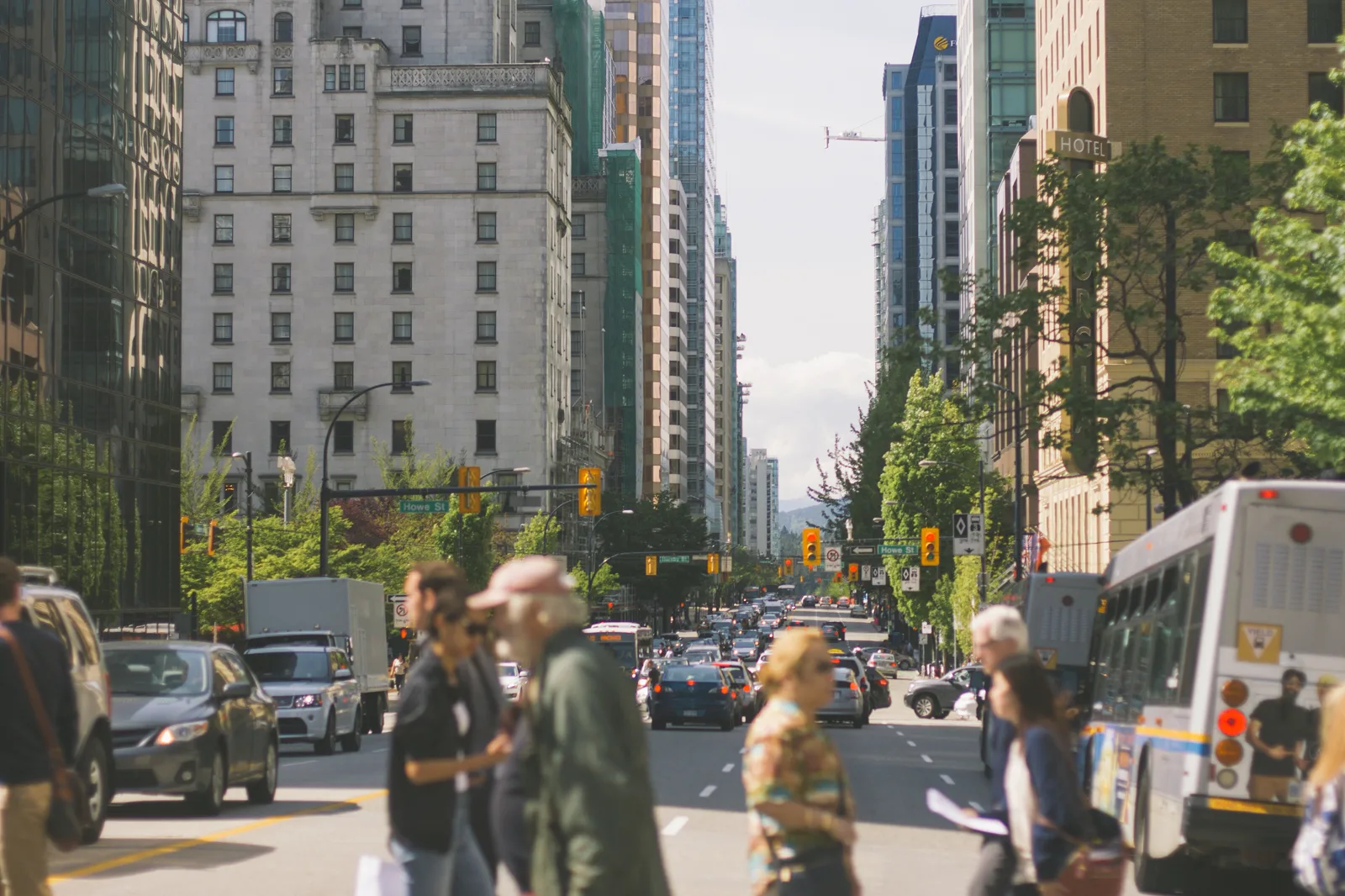 Pedestrians, out of focus in the foreground, cross a downtown street lined with high-rise buildings and trees while a city bus, cars and a truck wait at traffic lights