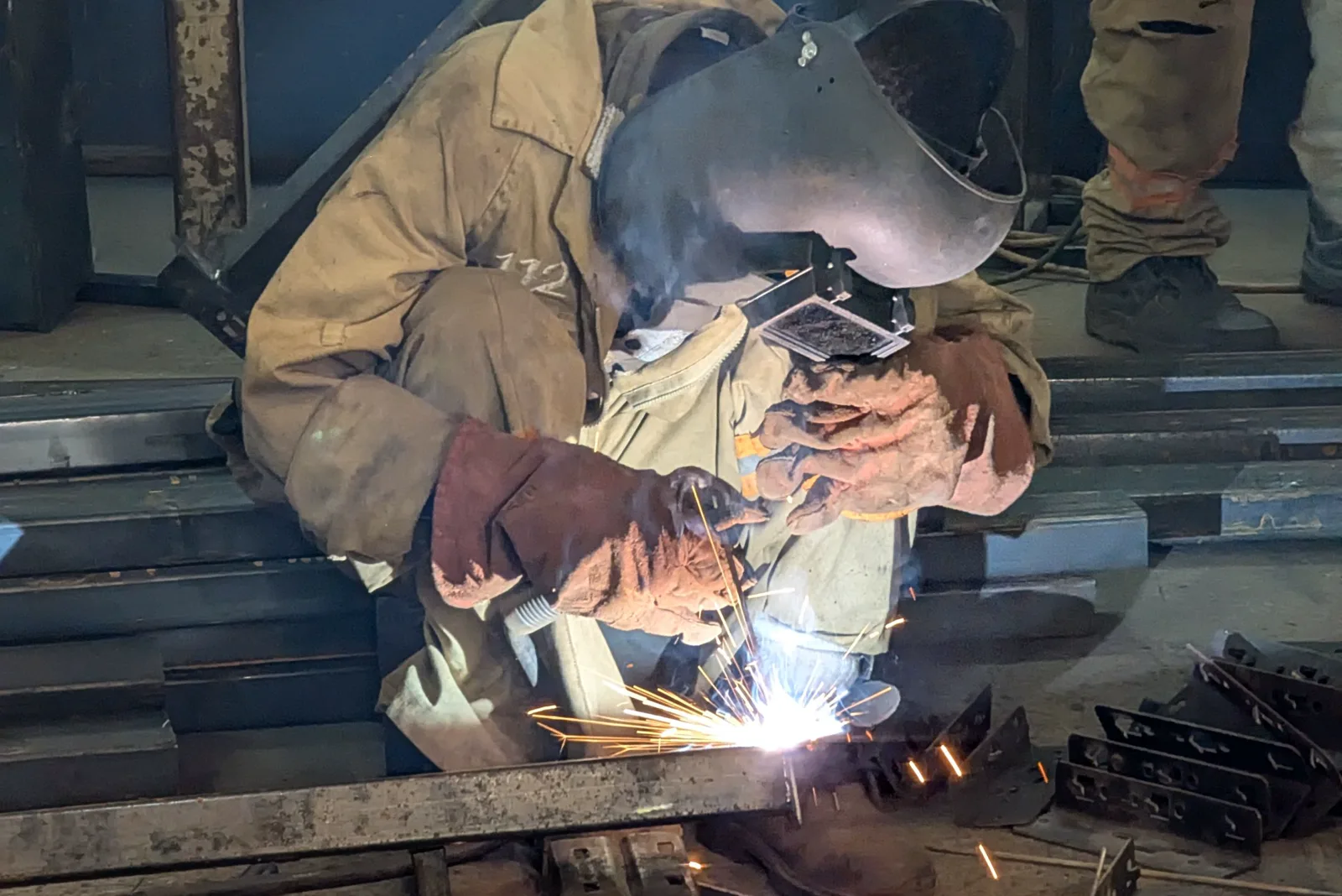 A worker in a welding helmet, leather gloves and a tan jacket crouches over a steel beam as sparks fly from the weld