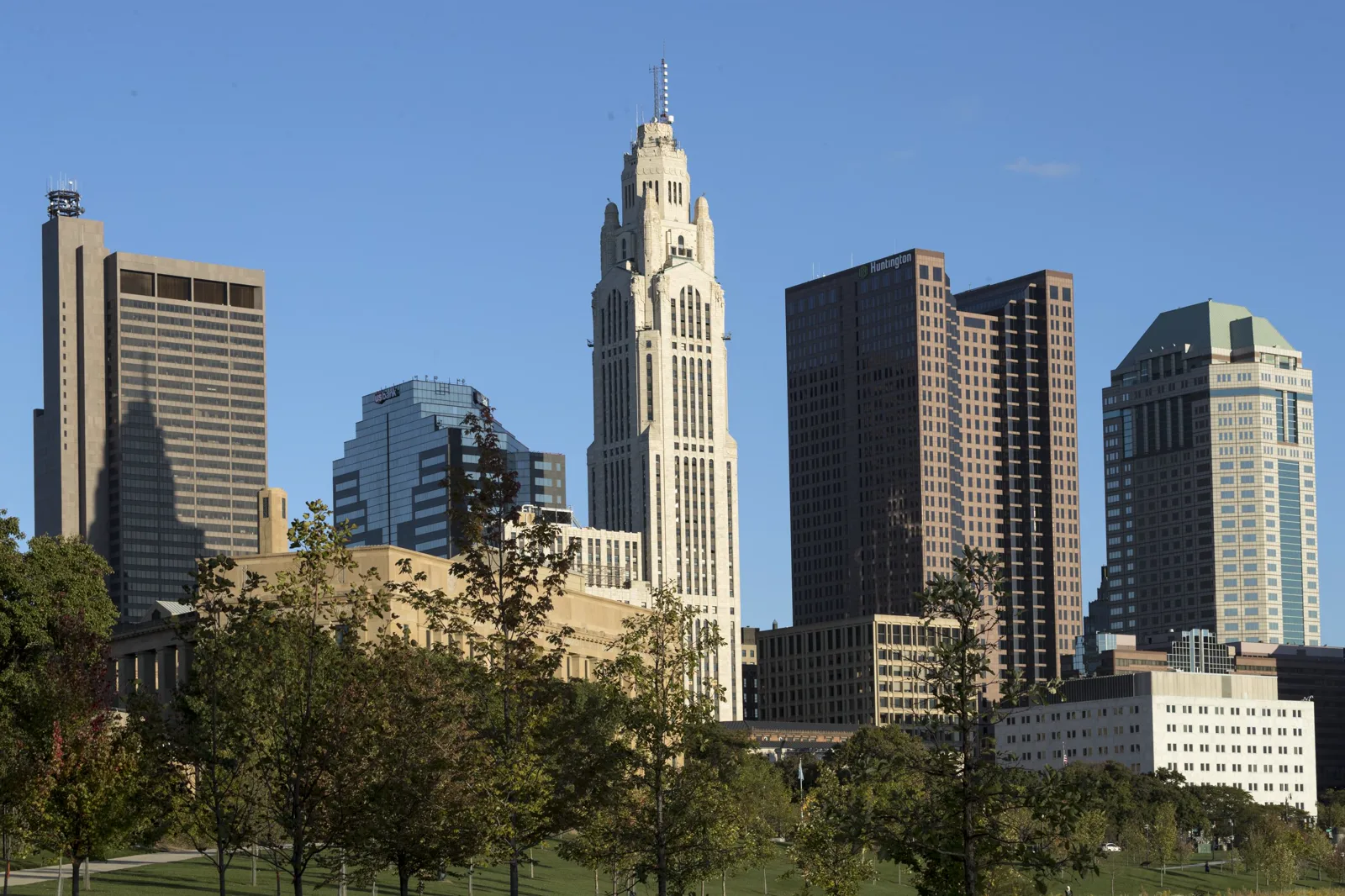 Downtown office towers under a clear blue sky, with a white stepped Art Deco tower in the middle and a row of green trees and lawn in front