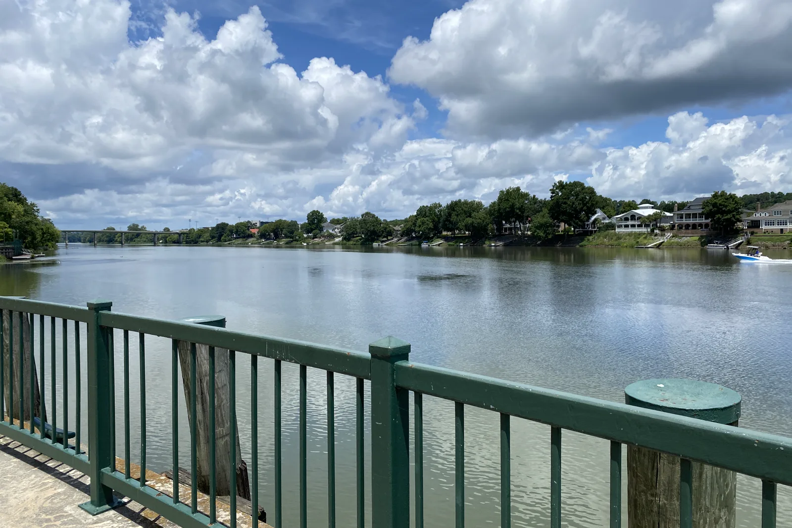 A wide, calm river under a sky of white clouds, seen over a green metal railing, with trees and large houses along the far bank and a road bridge in the distance