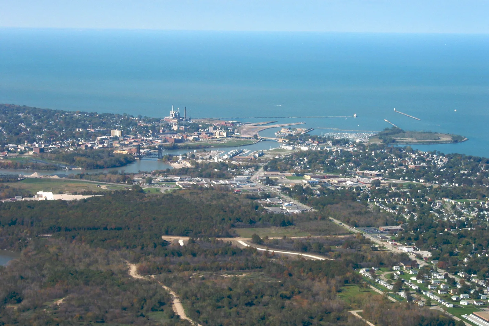 An aerial view of a lakeside city where a river meets a harbor with breakwaters, a lighthouse and a marina, with a power plant's smokestacks, rows of houses and woods in the foreground and a wide lake reaching the horizon