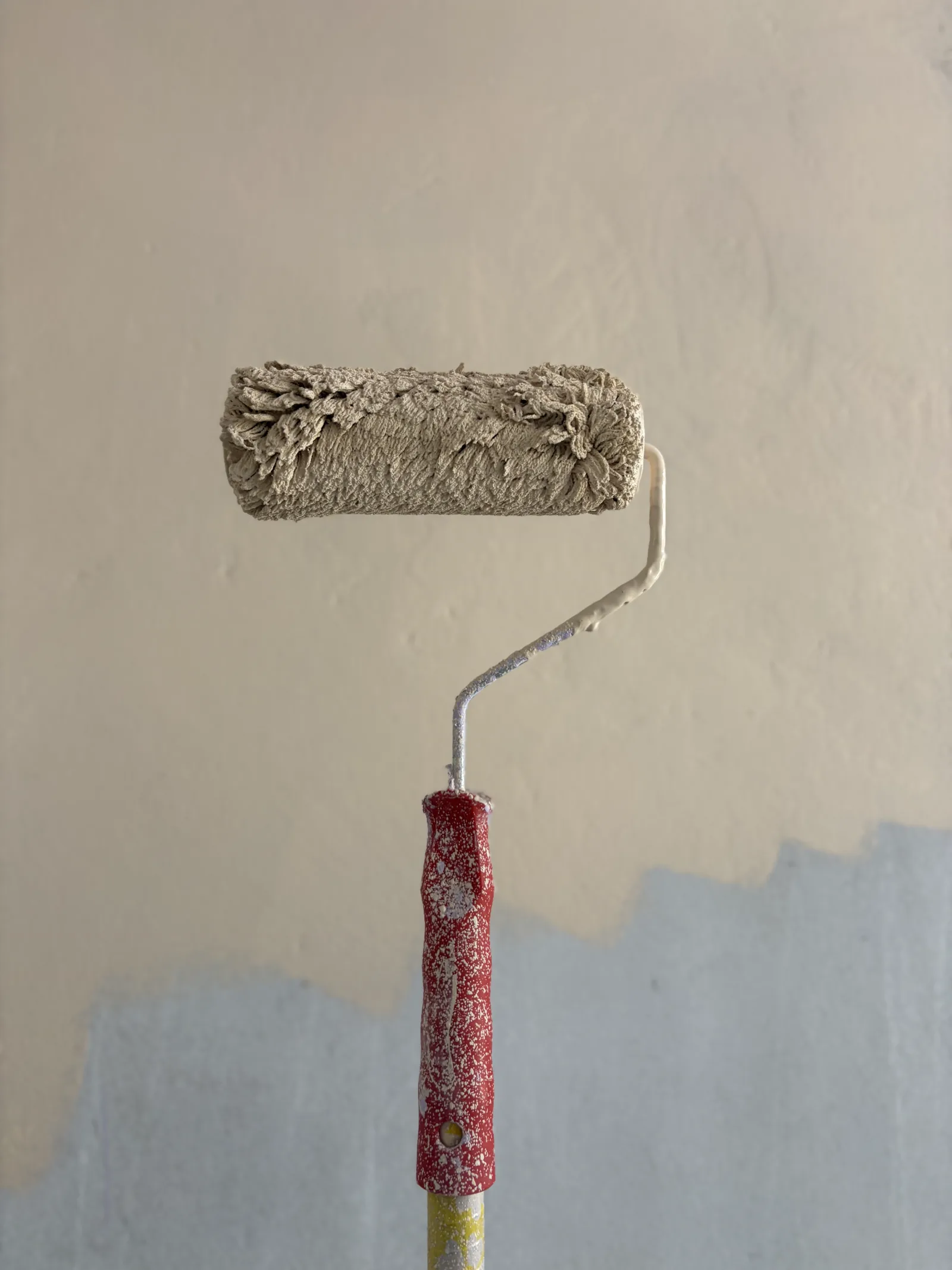A paint roller with a red handle, coated in beige paint, held up against a wall that is beige above and pale blue below where the new coat has not reached