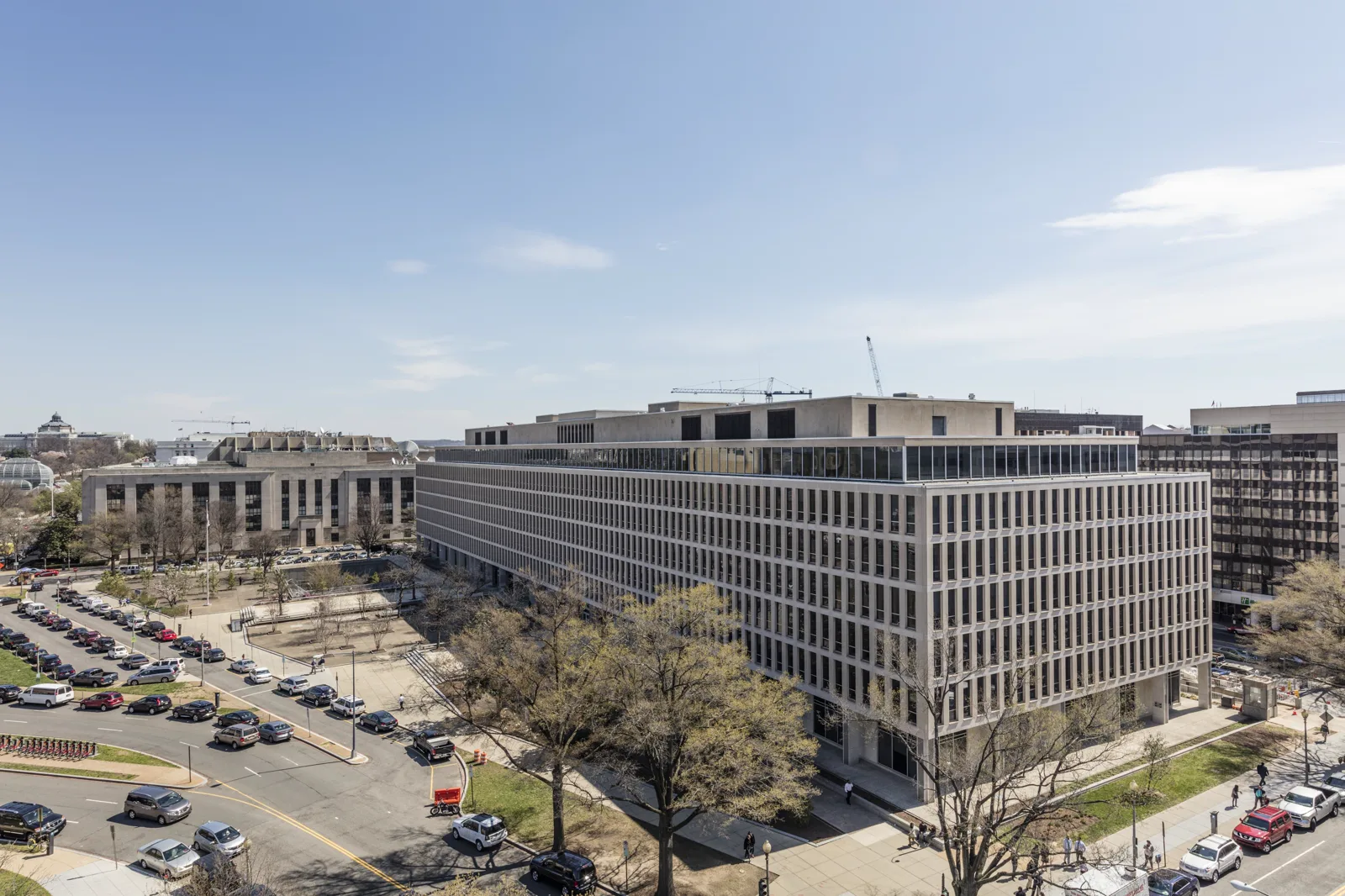 A long, pale six-story office building with rows of narrow vertical windows, seen from above, with a paved plaza, bare spring trees and a street lined with parked cars in front under a blue sky