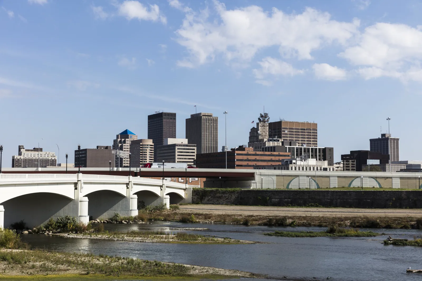Downtown office towers under a blue sky with white clouds, seen across a shallow river with grassy gravel bars and a white arched concrete bridge on the left