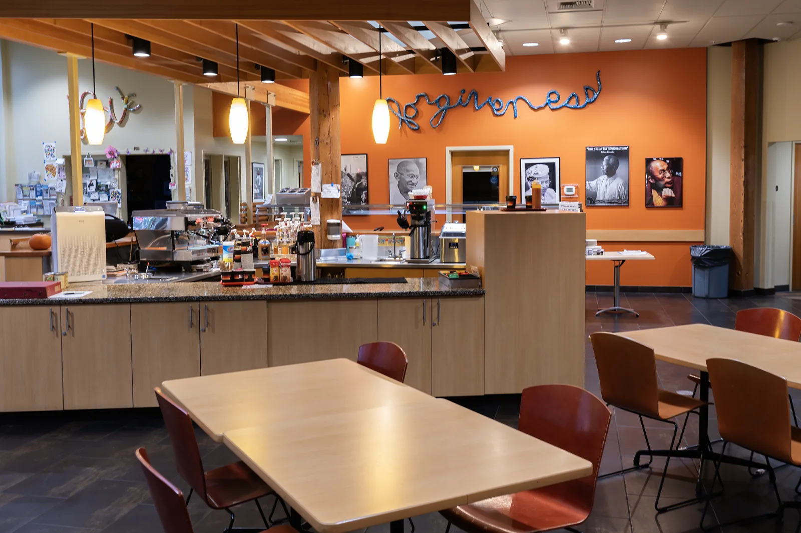 An empty cafe in a community center, with a long wooden counter holding a coffee urn, syrups and an espresso machine, wooden tables and red chairs in the foreground, and an orange wall behind with the word forgiveness in large blue script above framed black and white portraits
