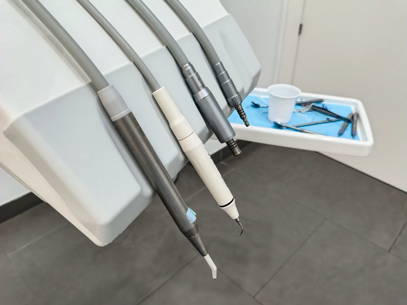 Dental handpieces and a scaler hang from the arm of a dental chair, with a tray of instruments and a plastic cup on a blue liner behind them