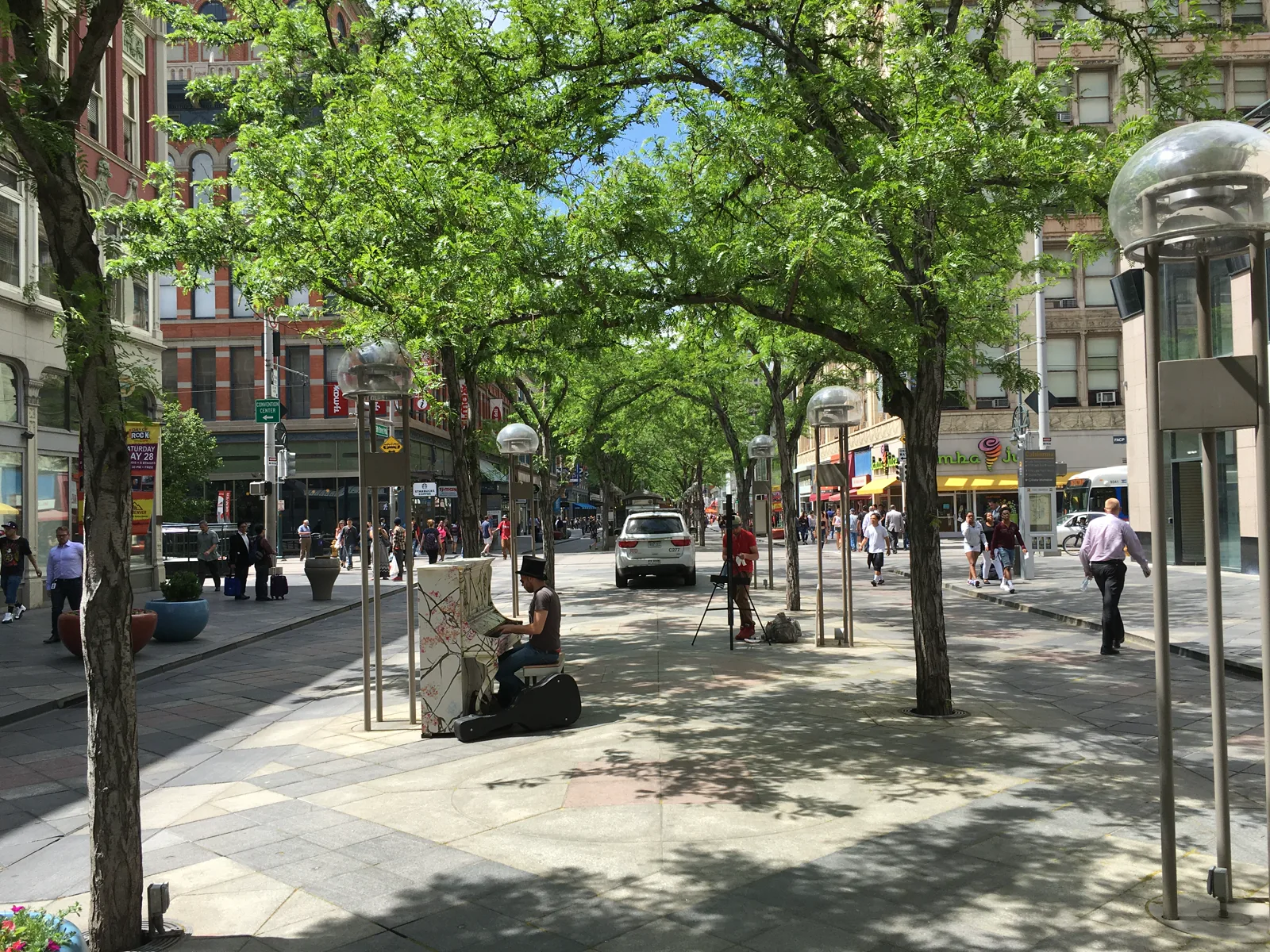 A tree-lined pedestrian street in downtown Denver with people walking at a distance, a man playing a painted street piano and a white SUV parked under the trees