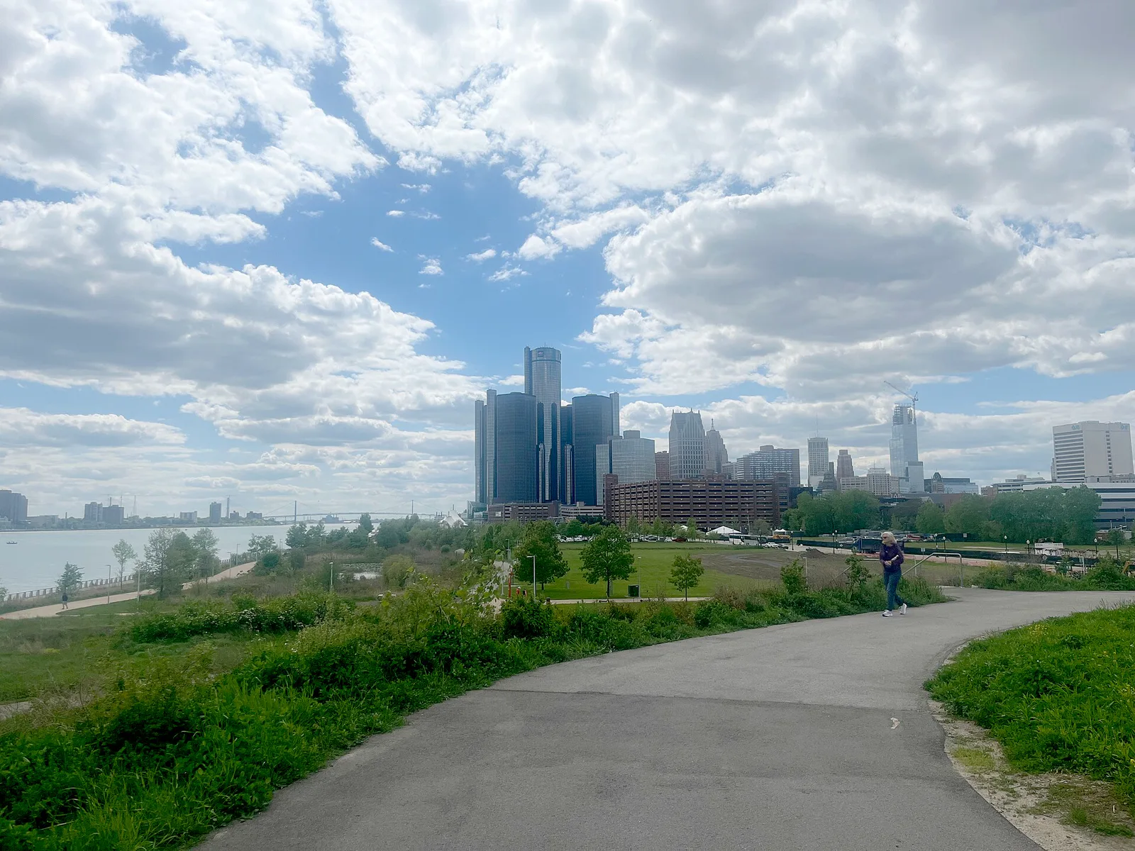 A paved path curves through grass and shrubs on the Detroit riverfront toward the dark glass towers of the Renaissance Center, with the Detroit River on the left, open lawns and parking in the middle distance and a single walker on the path