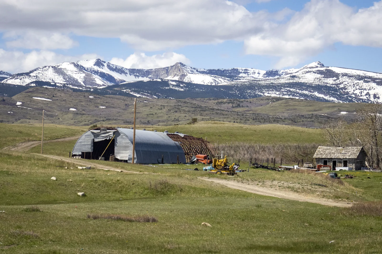 A metal quonset barn with part of its roof torn away, farm machinery and an old wooden cabin on green grassland, with snow-covered mountains behind