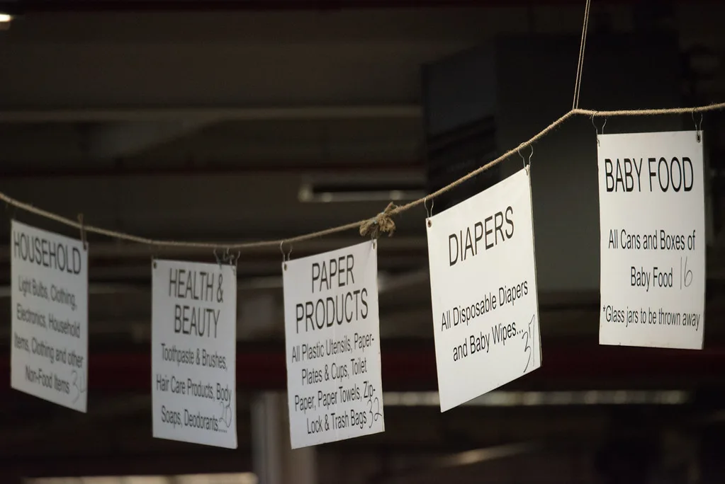 Hand-lettered signs hanging from a rope in a dim warehouse mark sorting stations for household goods, health and beauty items, paper products, diapers and baby food