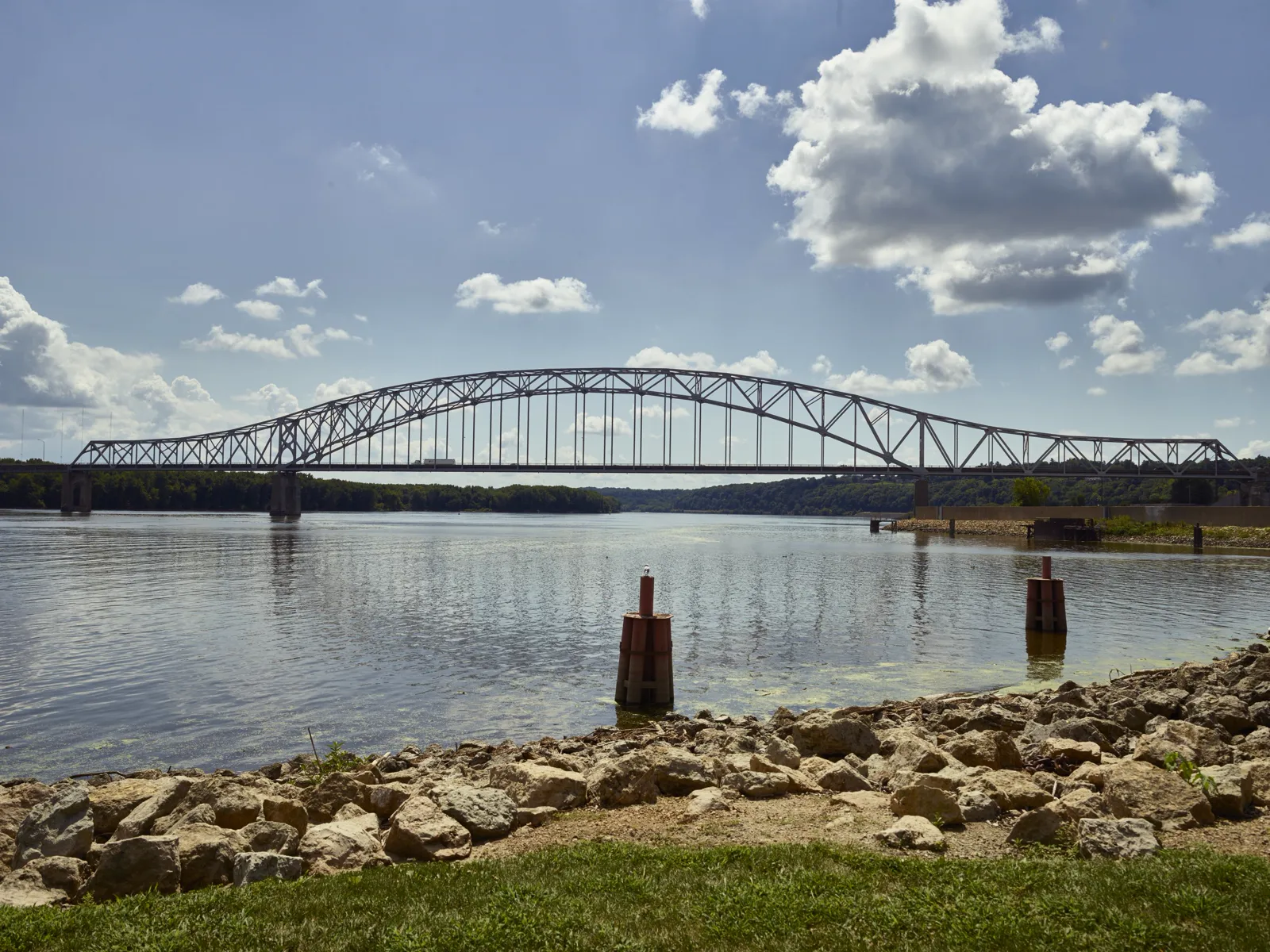 A long steel arch bridge spans a wide river under a blue sky with large white clouds, seen from a rocky bank with grass in the foreground and two red mooring posts in the water