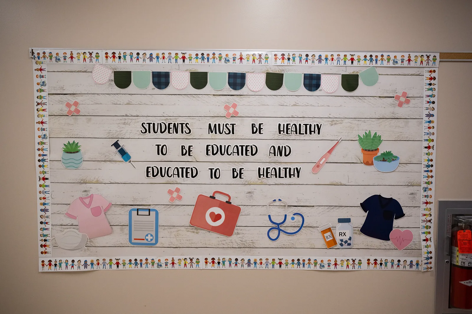 A bulletin board on a school wall reads Students must be healthy to be educated and educated to be healthy, decorated with paper cutouts of a stethoscope, a first aid kit, a thermometer, a syringe, a pill bottle, plants and a heart