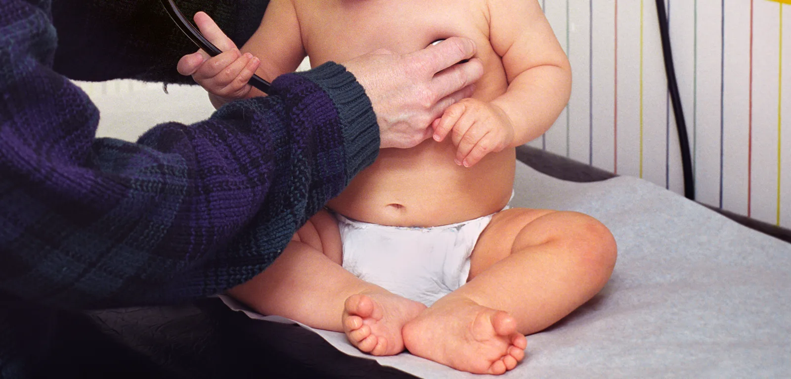 A physician in a dark plaid sweater holds a stethoscope to the chest of an infant in a diaper sitting on a paper-covered exam table, the baby's hands resting on the doctor's hand