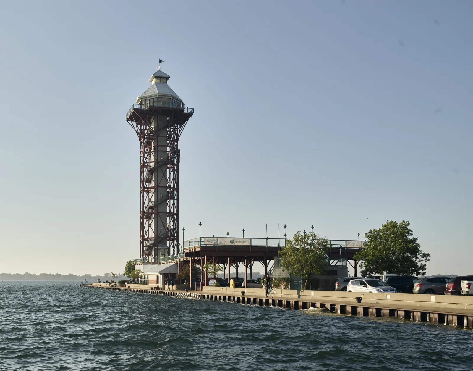 A tall observation tower with a red steel frame and a white top stands at the end of a concrete pier on choppy water, beside a raised deck, trees and a row of parked cars