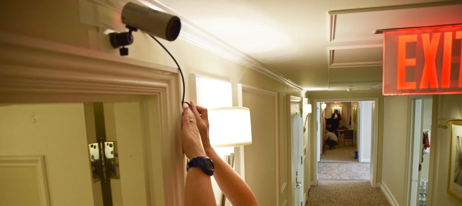 Two raised arms, one with a dark blue wristwatch, fasten the cable of a small security camera mounted above a doorway in a carpeted corridor with wall lamps and a red exit sign