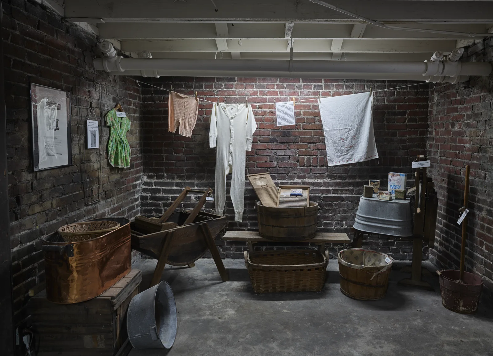 A brick cellar room set up as a museum display of washday, with copper and wooden tubs, baskets, a washboard and long underwear hanging on a line, each object with a small label