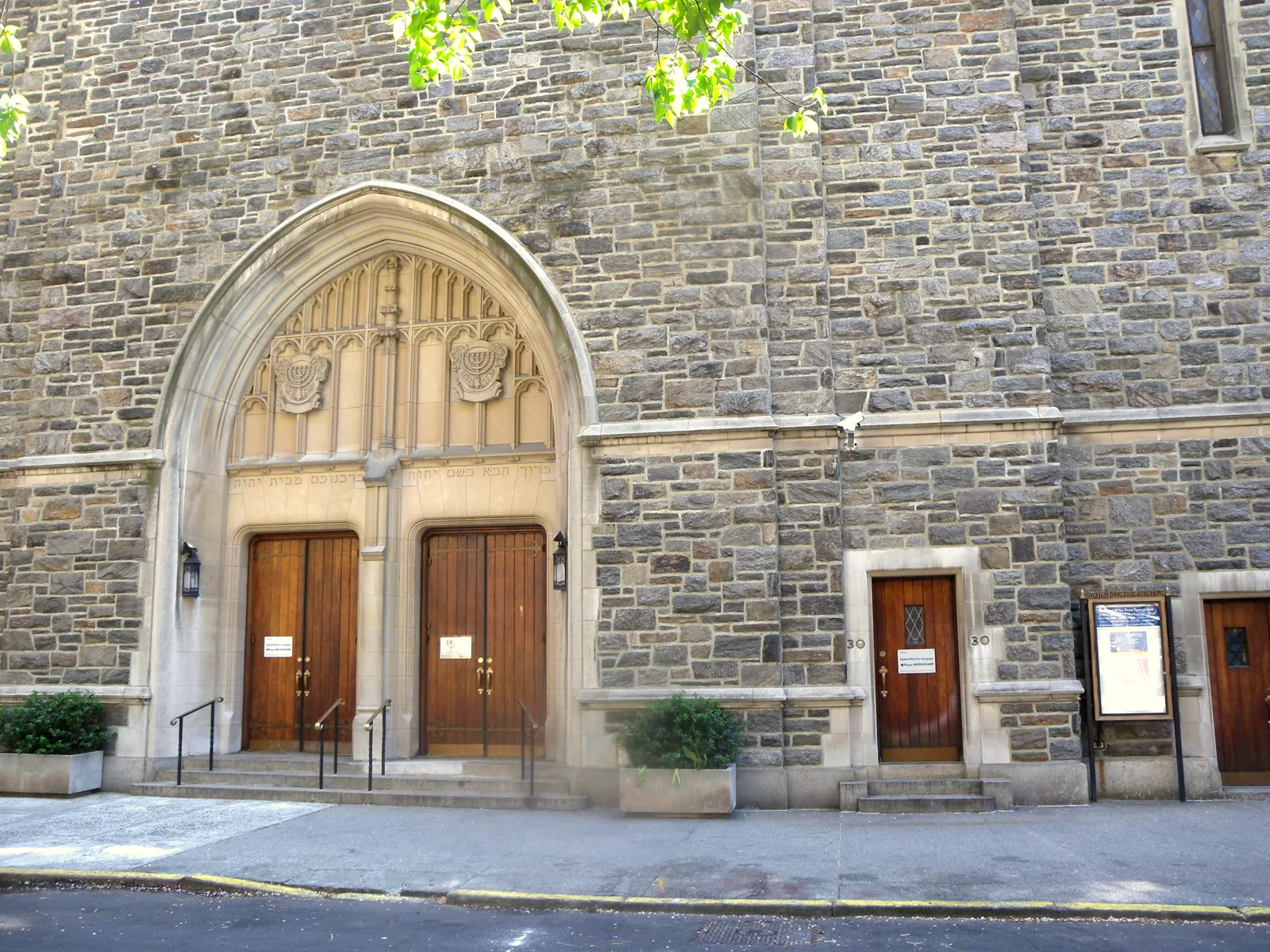 The stone front of a synagogue with a pointed stone arch over two tall wooden doors, a white security camera mounted on the wall beside it and a smaller side door with a notice board