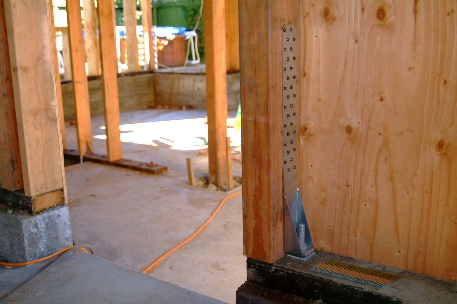 A perforated metal strap and bracket bolted to the foot of a wooden wall post where it meets the foundation sill, with bare wall framing and a concrete floor behind it