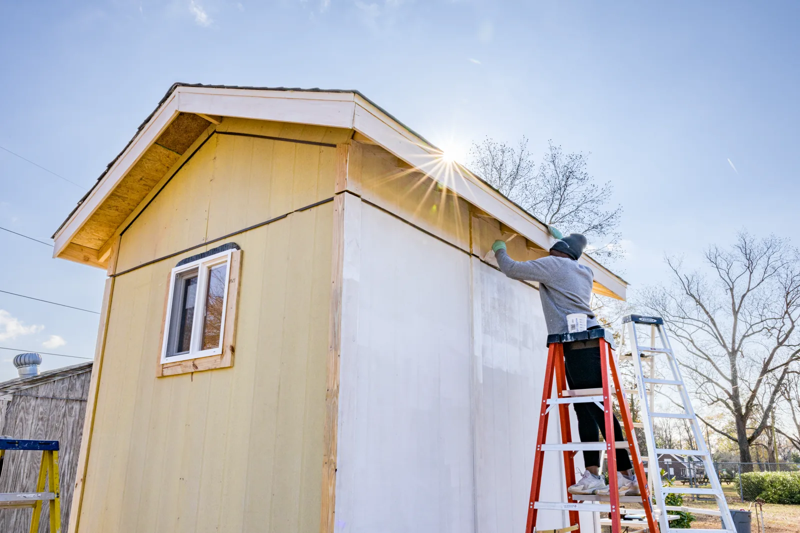 A person in a gray sweatshirt and knit cap, seen from behind on an orange stepladder, paints the white trim of a small pale yellow wooden building under a low sun, with a second ladder beside it
