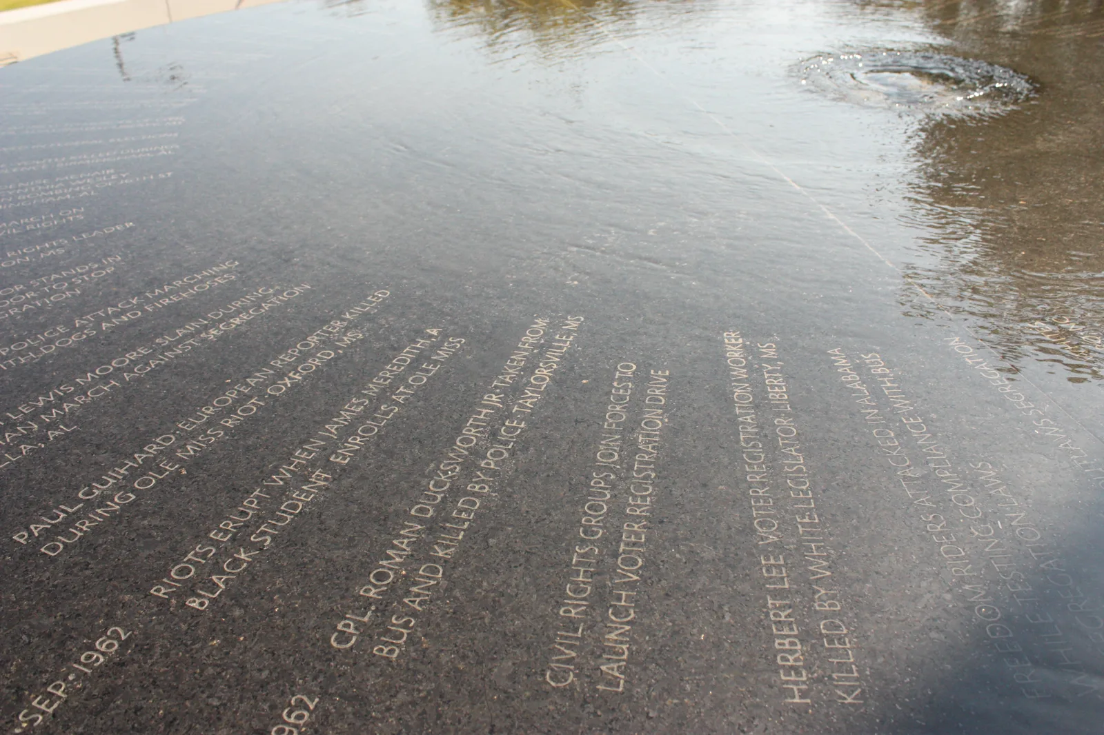 Water runs in a thin sheet over a round black granite table carved with dated lines of text, with a small upwelling of water at the top right and a reflection of trees on the wet stone